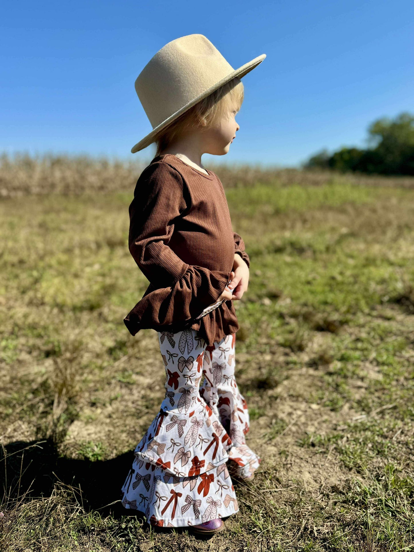 Child wearing a brown top and patterned pants with a wide-brimmed hat in a grassy field.